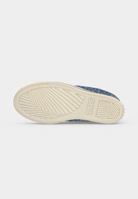 Semelle de chaussure avec une bande de roulement en caoutchouc texturé beige présentant des rainures diagonales et le nom de la marque "GUESS" embossé près du talon.