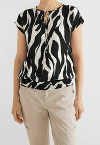 Schwarze und weiße Kurzarmbluse mit Zebra-Print, ausgestattet mit einem Schlüsselloch-Ausschnitt und einem elastischen, gerafften Saum. Kombiniert mit beigen Hosen.