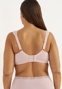 Frau mit langen braunen Haaren, die einen verstellbaren, hellrosa BH und passende Unterwäsche mit Spitzenbesatz trägt, von hinten gezeigt.