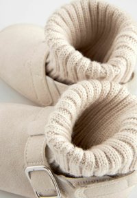 Beežid suede saapad ribatud kudumisega äärtega, küljes rihm ja metallne pandlaga. Sile tekstuur koos ümarate varvaste kujundusega.