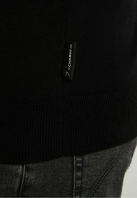 Ourlet de pull en tricot noir avec une petite étiquette noire en tissu portant la mention « CHASIN' » et un jean en denim gris foncé en dessous.