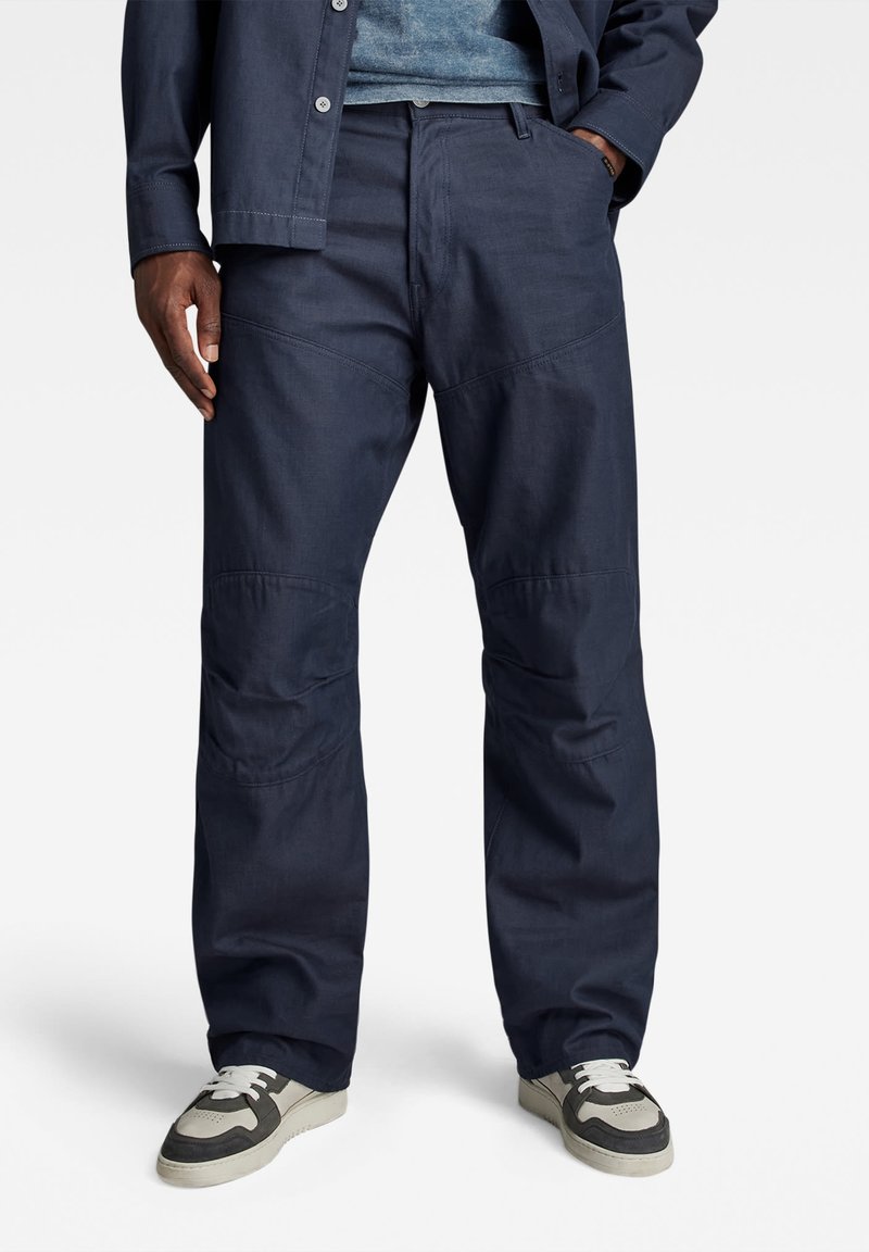 Marineblauwe werkbroek van textuurstof; met stikselaccenten en een ontspannen pasvorm. Gepaard met grijze en witte sneakers.
