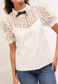 Weiße Bluse aus floraler Spitze mit Puffärmeln, rüschigem Ausschnitt und schwarzem Schleifenakzent. Strukturierter Stoff und mehrlagiges Design verleihen Tiefe.