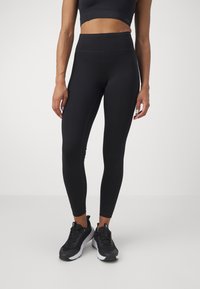 Des leggings noirs taille haute en matériau extensible, avec une texture lisse et une coupe ajustée, associés à des chaussures de sport noires.
