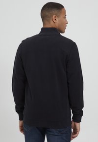 Svart fleecejakke med høy krage, med en glatt tekstur og ribbede mansjetter, vist fra baksiden med et minimalistisk design.
