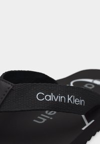 Μαύρο σαγιονάρι Calvin Klein με υφή στο λουράκι που φέρει το όνομα της μάρκας με λευκά γράμματα.
