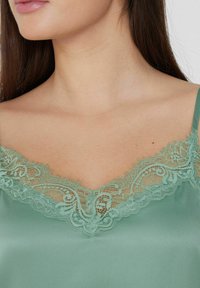 Camisole en satin vert émeraude avec une délicate dentelle le long d'un décolleté en V, avec une texture lisse et des bretelles ajustables.