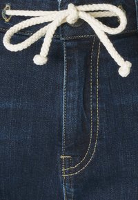 Tummat farkut, joissa on punottu valkoinen köysi nyörinä vyötäröllä, ja kontrastina keltaista tikkausta saumoissa.