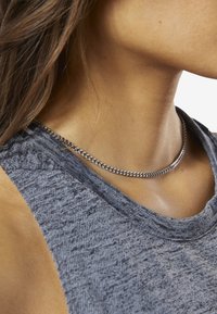 Silverkedjan ligger runt en hals, med en slät, rundad design och sammanlänkade länkar, bärs över en texturerad grå tyg.