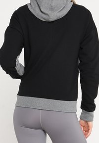 Haut à capuche noir et gris avec une texture douce, présentant une capuche et des poignets gris contrastés, ainsi qu'un ourlet à côtes. Design simple et sportif.