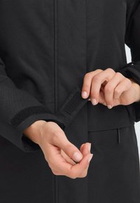 Svart jacka med en texturerad tyg, synliga kardborrestängningar vid handleden och en snygg, strukturerad design. Händer justerar ärmslutet.