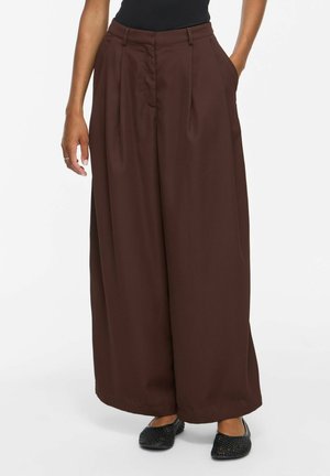 Personne portant un pantalon large marron foncé avec des poches et des chaussures plates tissées noires, debout devant un fond uni.