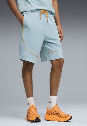 Mężczyzna w jasnoniebieskich szortach z pomarańczowym obszyciem, białych skarpetkach Puma i jaskrawo pomarańczowych butach do biegania Puma na szarym tle gradientowym.