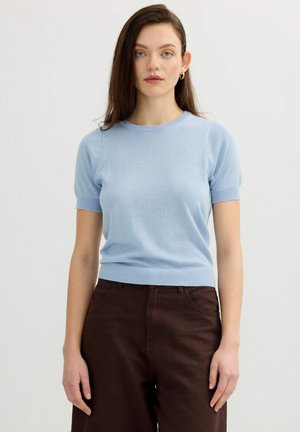 Mujer con cabello largo y castaño que lleva un top de punto azul claro de manga corta y pantalones marrón oscuro, de pie frente a un fondo liso.