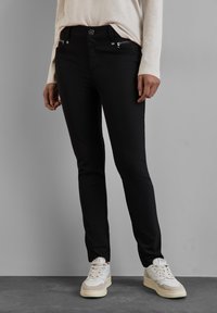 Černé slim-fit džíny s předními kapsami a stříbrným kováním, v kombinaci s bílými teniskami. Hladká textura, minimalistický design.