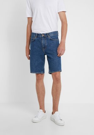 Szorty dżinsowe w średnim niebieskim kolorze, charakteryzujące się klasycznym krojem, bocznymi kieszeniami i szlufkami na pasek. W połączeniu z gładką białą koszulką i białymi trampkami.