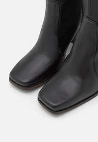 Bottines en cuir noir avec une finition brillante, design à bout carré et détails de couture visibles. Présente un talon empilé pour l'élévation.