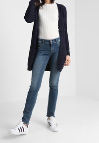 Námořnický pletený kardigan přes bílý žebrovaný svetr v kombinaci s modrými úzkými džínami a bílými teniskami s černými pruhy.