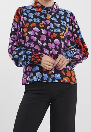 Femme portant une blouse noire à manches longues avec des motifs floraux colorés en bleu, violet, rouge et orange, associée à un pantalon noir.
