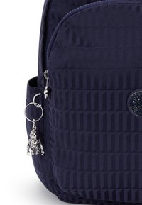 Granatowy plecak o teksturowanej powierzchni, z okrągłym logo i srebrnym breloczkiem w kształcie robota przymocowanym do suwaka.