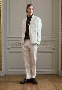Vit kavaj och byxor av lättviktsmaterial, tillsammans med en svart skjorta med prickar. Glansiga svarta skor fulländar outfiten.