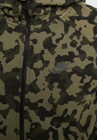 Mikina s kapucí na zip v maskáčovém vzoru s kombinací tmavě a světlounce zelených odstínů, texturovanou tkaninou a jemným logem Nike na hrudi.