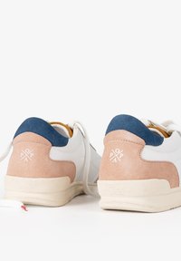 Zapatilla blanca con acentos de ante rosa y un parche en la parte trasera de color azul marino. Presenta una suela texturizada color crema y un logotipo bordado en la parte trasera.