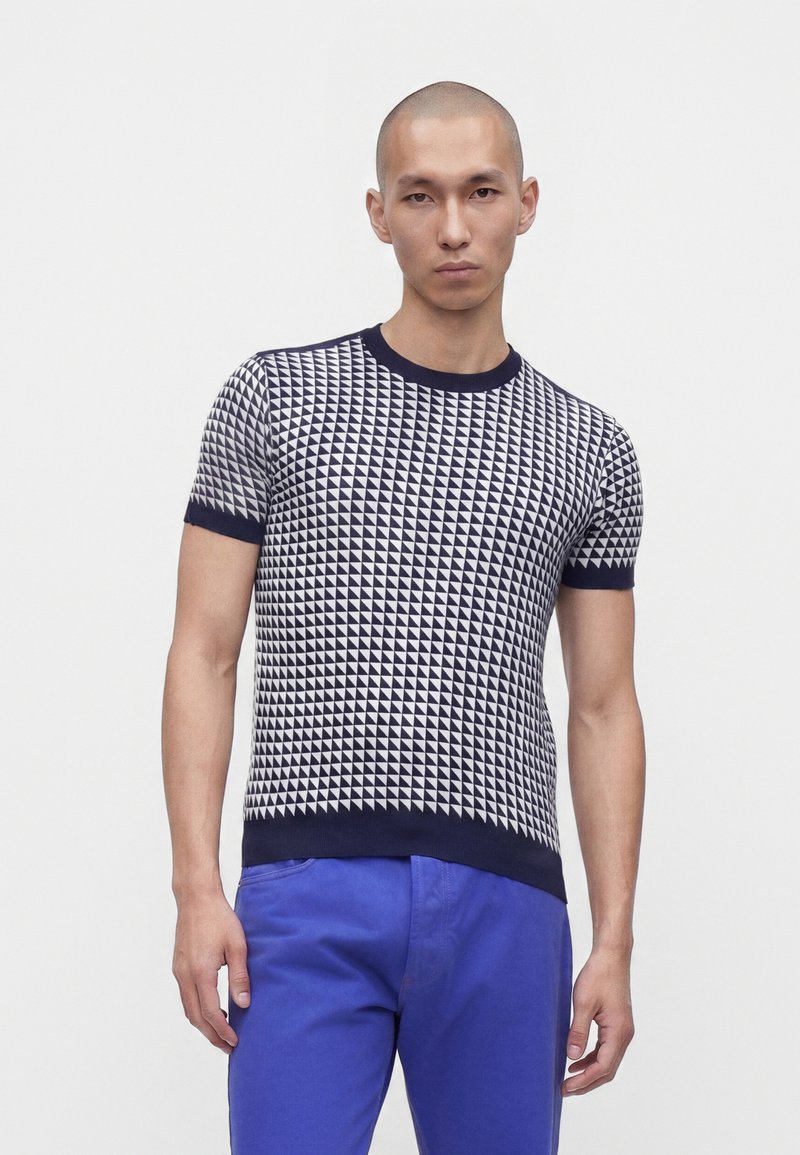 Homme portant une chemise à manches courtes à motif géométrique bleu marine et blanc avec un pantalon bleu, debout devant un fond blanc uni.