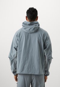 Ανοιχτό γκρι μπουφάν με κουκούλα, διαθέτοντας ελαστικούς μανσέτες και χαλαρή εφαρμογή. Ματ υφή με απλό σχέδιο, χωρίς ορατά μοτίβα ή λογότυπα.