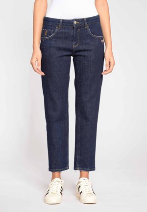 Nainen, jolla on yllään tummansiniset suoralahkeiset farkut, joissa on rispaantuneet lahkeet, sekä valkoiset tennarit mustilla raidoilla, seisoo yksiväristä taustaa vasten.