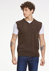 Brun stickad väst med V-ringning och ribbad fåll, bärs över en vit T-shirt. Modellen har matchat med blå denimjeans.