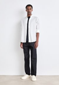 Camisa de lino blanca sobre una camiseta negra, combinada con jeans de mezclilla oscura y zapatillas blancas. El ajuste es relajado con un cierre de botones en la parte delantera.