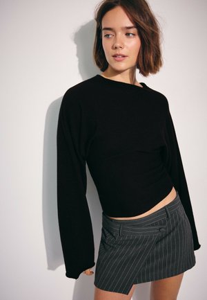 Jeune femme aux cheveux courts et bruns portant un haut noir à manches longues et une mini-jupe grise à fines rayures, regardant sur le côté sur un fond blanc.