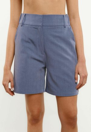 Magas derekú, szabott kék rövidnadrág övbújtatókkal, elülső cipzárral és oldalzsebekkel, egy fehér háttér előtt álló személyen viselve.