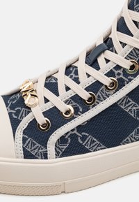 Gros plan d'une basket marine avec des lacets blancs, des œillets dorés, un tissu à motifs, une semelle beige et un charm logo MK doré sur les lacets.
