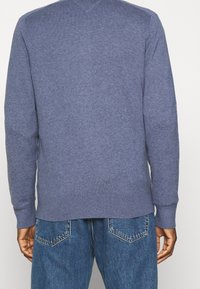 Maglione blu a maglia con maniche lunghe e scollo a V, con polsini e orlo a coste. Indossato sopra jeans di denim blu con cuciture delle tasche posteriori visibili.