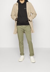 Abrigo trench beige sobre una sudadera negra, combinado con pantalones verde oliva y zapatillas blancas. Diseño informal, corte estándar con puños remangados.