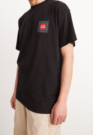 T-shirt in cotone nero con tasca quadrata sul petto decorata da un'icona rossa bordata da un motivo verde e nero. Maniche corte, vestibilità rilassata.