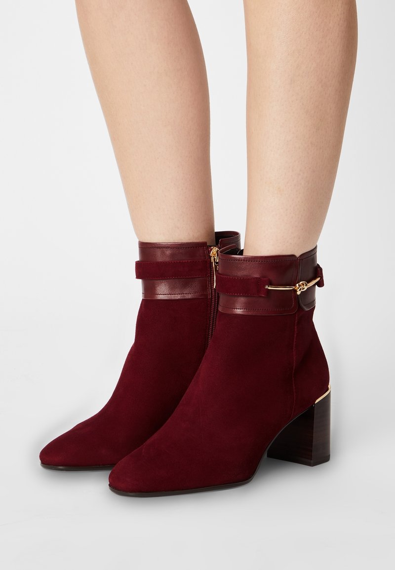 Bottes en daim bordeaux montant à la cheville avec talons blocs, garniture en cuir, détail de boucle dorée, portées sur jambes nues devant un fond uni.