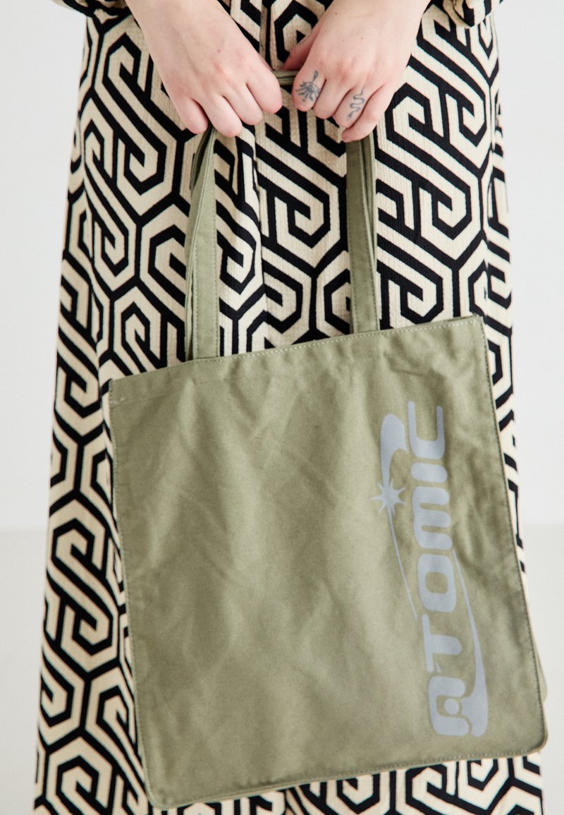 Sac fourre-tout en toile verte avec doubles poignées, arborant un logo "Atomic" gris bien visible et une texture douce ; robe à motifs en arrière-plan.