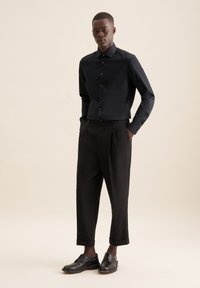 Camicia nera con bottoni, pantaloni neri su misura con pieghe e scarpe di pelle nera. Texture liscia, design elegante e silhouette aderente.