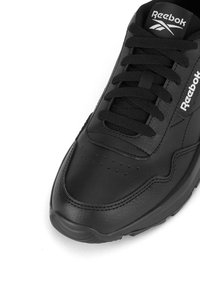 Juodos odos sportinio stiliaus batai su lygiu paviršiumi, apvaliu priekiniu galu, raišteliais, tekstūrinėmis detalėmis ir logotipu ant liežuvėlio.