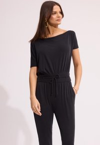 Czarny kombinezon z krótkimi rękawami, elastycznym pasem, szczegółami z troczkiem i bocznymi kieszeniami. Gładka tekstura i luźny krój.