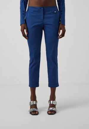 Personne portant un pantalon bleu slim court avec fermeture bouton et zip, associé à des sandales argentées à bouts ouverts avec larges brides à boucles.