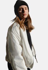 Weißer Bomberjacke mit glattem, glänzendem Finish, ausgestattet mit einem Reißverschluss und gerippten Bündchen, über einem schwarzen Pullover getragen und kombiniert mit einer schwarzen Strickmütze.