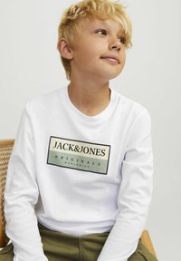 Ilga rankovė balta marškiniai iš Jack & Jones, su juodu ir žaliu stačiakampiu logotipu, turinčiu tekstą. Medvilnės audinys, laisvas fasonas.