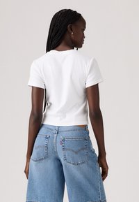 Tricou scurt, alb, cu mânecă scurtă, combinat cu pantaloni scurți din denim deschis, cu croi larg, având buzunare la spate și un etichetă vizibilă a brandului.