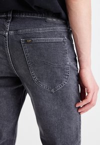 Gros plan sur une personne portant un jean Lee gris foncé, montrant la poche arrière et une partie de la main droite reposant sur le côté.