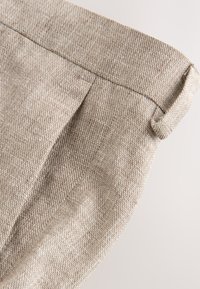 Close-up of beige and brown textured fabric pants with belt loop, sleek seam, and pleated detail on a light background.