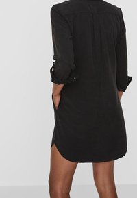 Black shirt dress with long sleeves, buttoned cuffs, and side pockets. Features a relaxed fit and curved hem. Made from lightweight fabric.